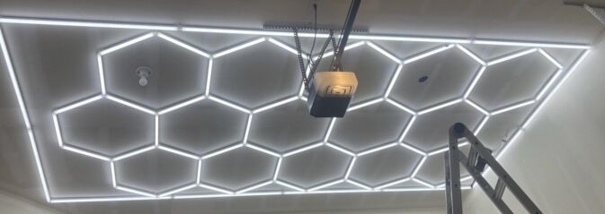 Hexagonal LED light panels are mounted on a ceiling near a garage door opener, with part of a ladder visible in the lower right corner.
