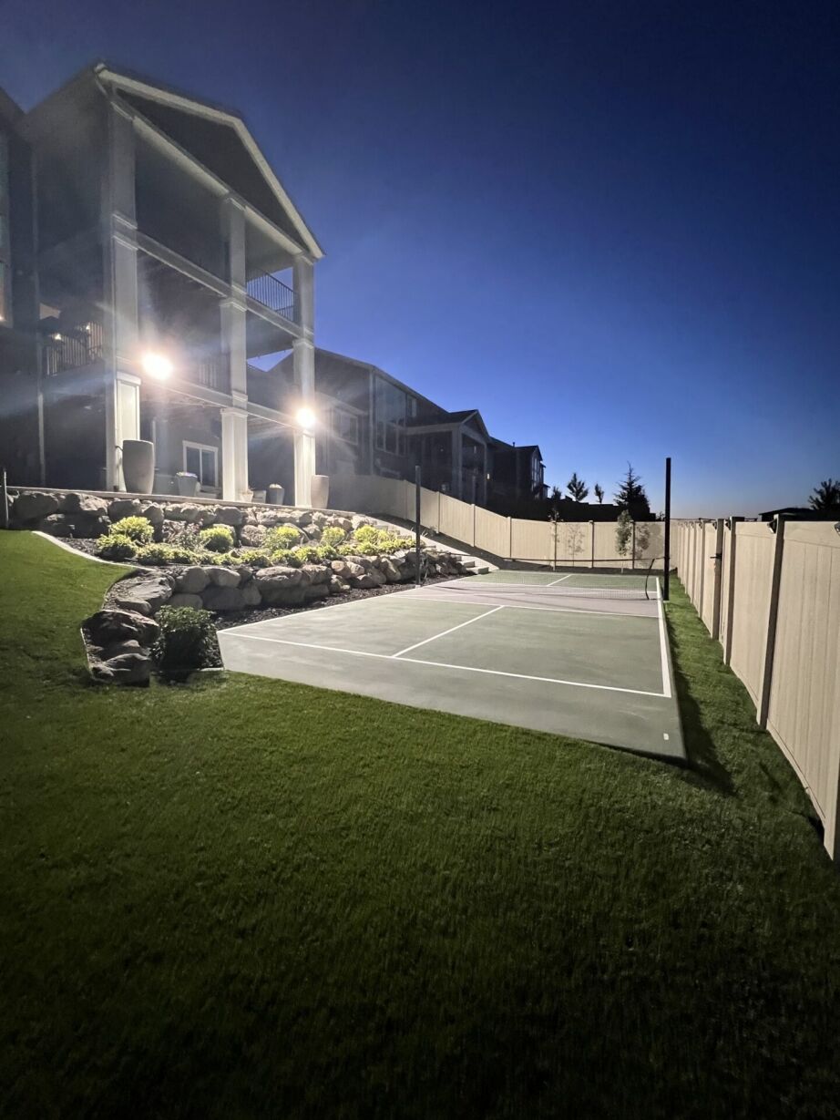 A lit pickleball court beside a modern house with landscaped rocks and plants, enclosed by a white fence, at dusk.