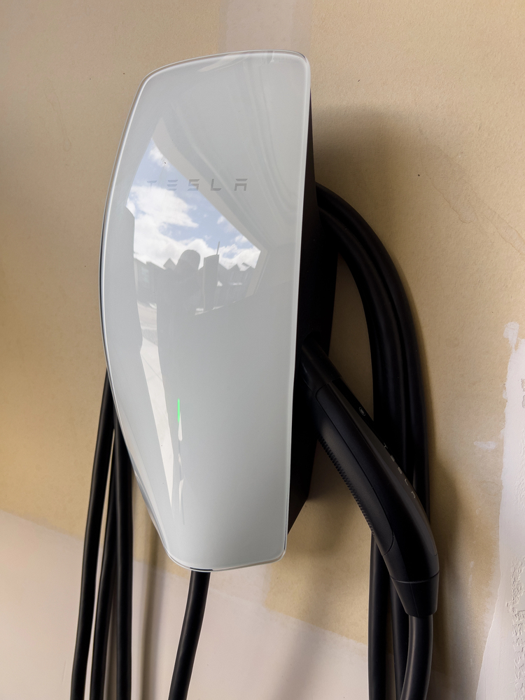 White Tesla wall connector with attached charging cable mounted on a beige wall, reflecting light and showing faint outdoor scenery.