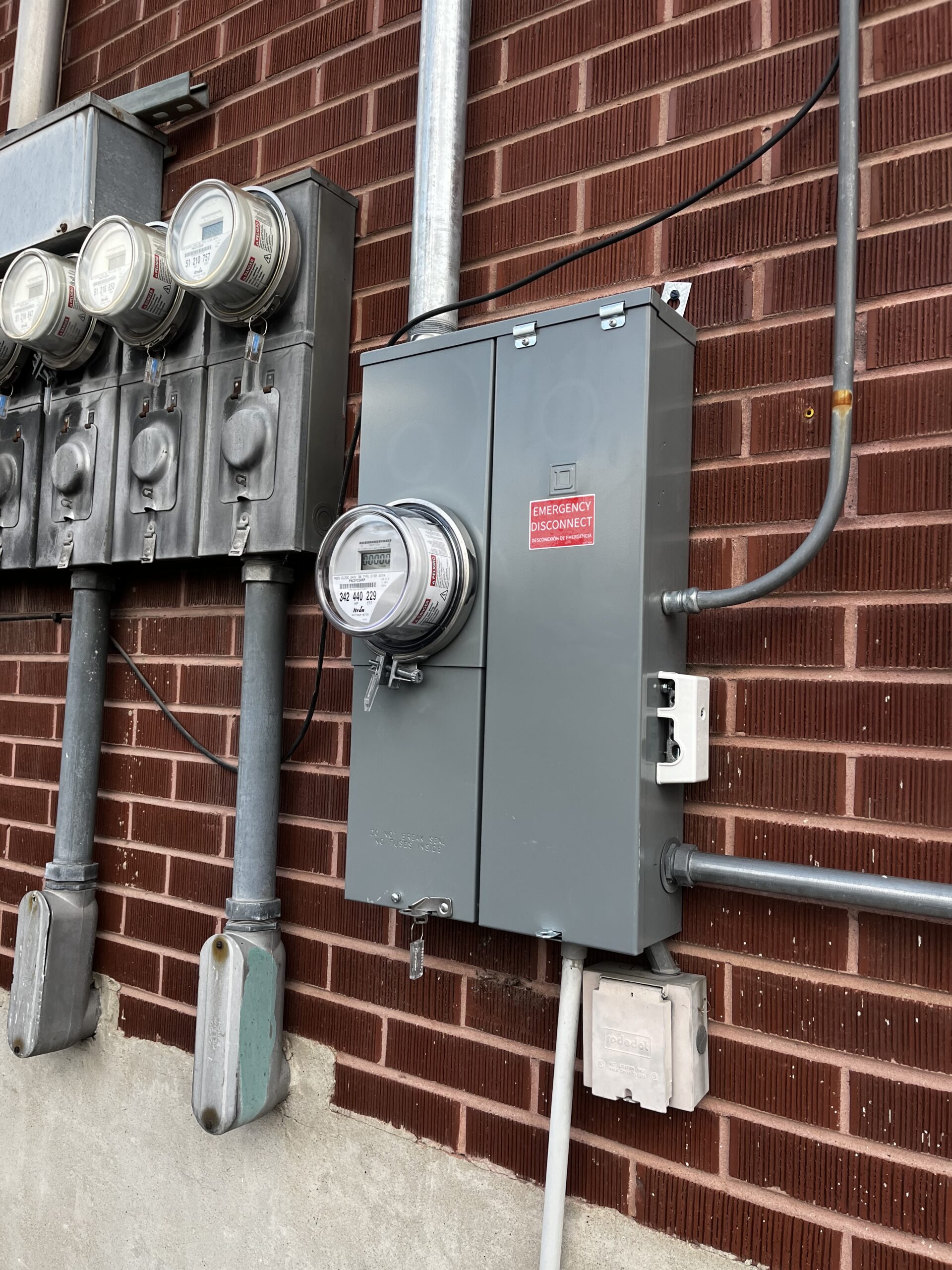 Several utility meters and electrical panels are mounted on a brick wall, with one labeled Emergency Disconnect and various pipes and cables connected.