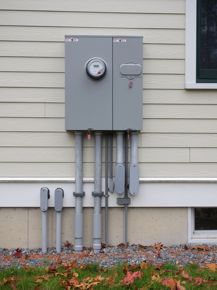 A gray electrical utility meter and breaker panel are mounted on the exterior siding of a house, with several conduit pipes running vertically below.