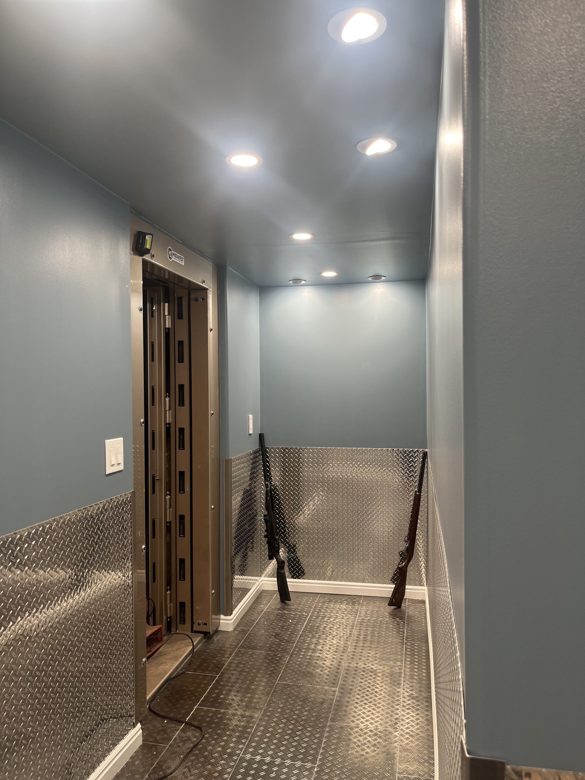 A secure room with blue walls, metal paneling, a reinforced door, ceiling lights, and two rifles propped against the wall.