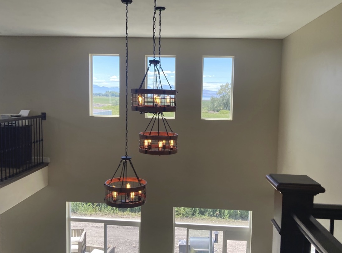 Three round, rustic-style pendant lights hang from the ceiling in a two-story room with large windows showing an outdoor landscape.