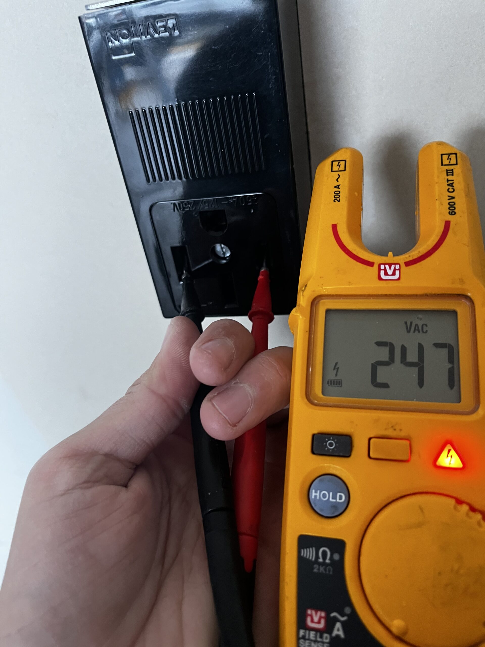 A hand holds two multimeter probes in a power outlet while the digital multimeter display reads 247 volts AC.