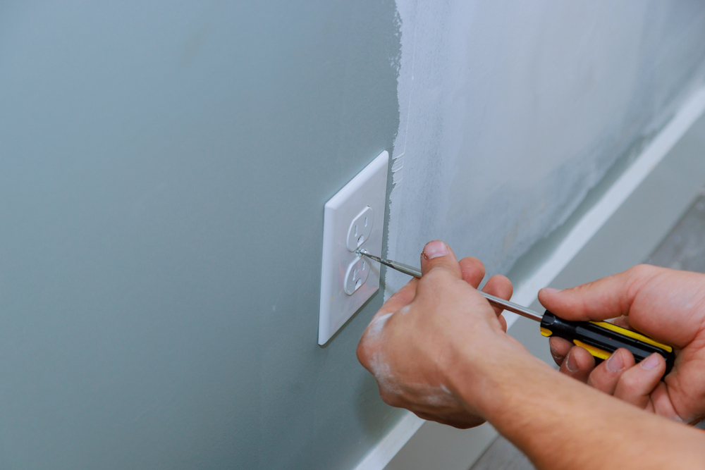 A person uses a screwdriver to install or remove a wall outlet cover on a light green painted wall.