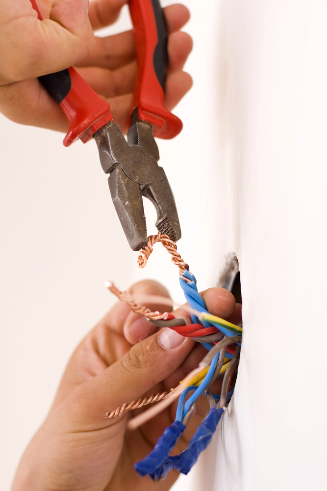 A person uses pliers to cut exposed copper wires protruding from a wall, with various colored wires bundled together and some ends wrapped in blue tape.