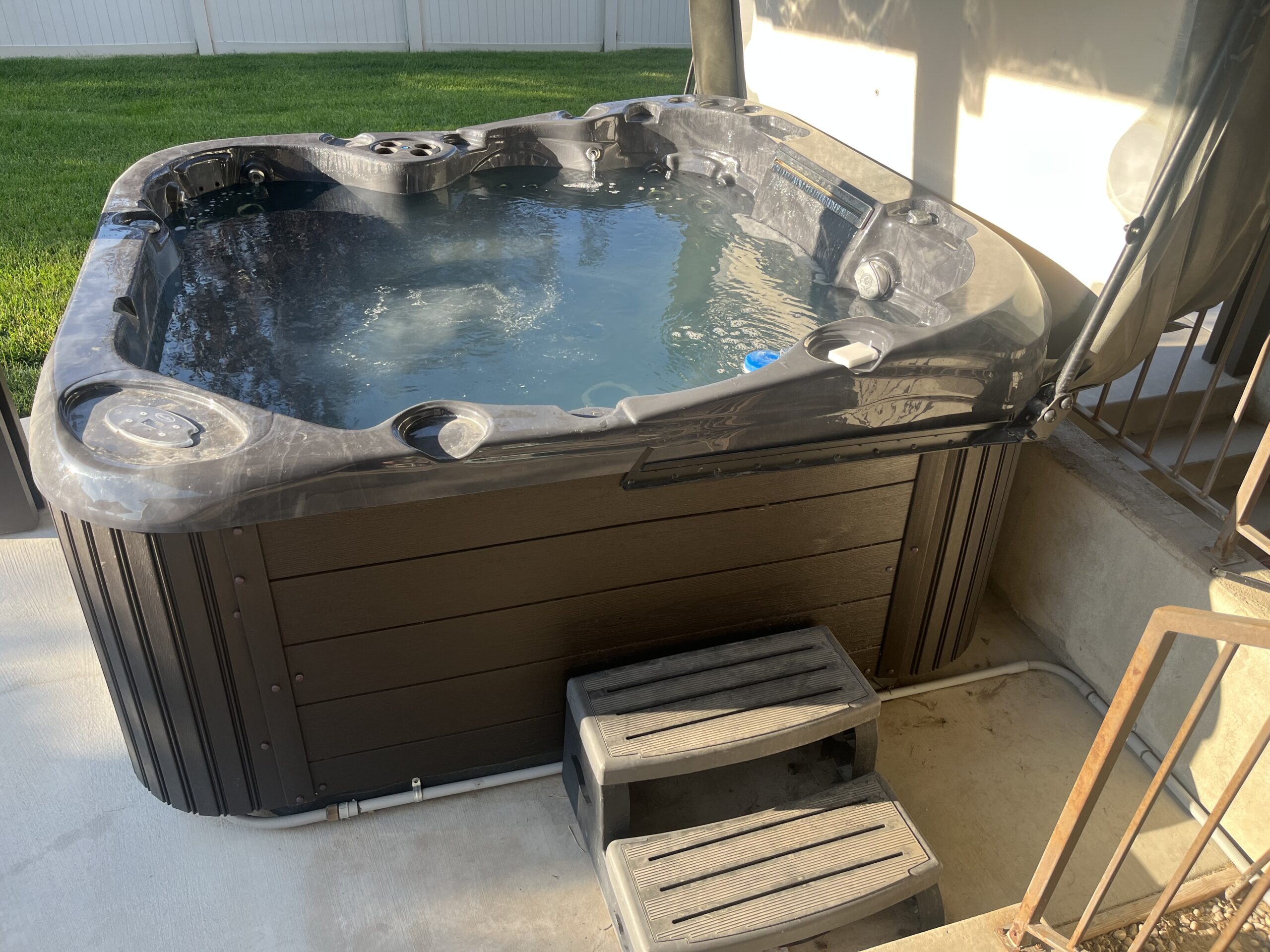 A black outdoor hot tub filled with water is situated on a concrete patio, with plastic steps in front and a grassy yard in the background.