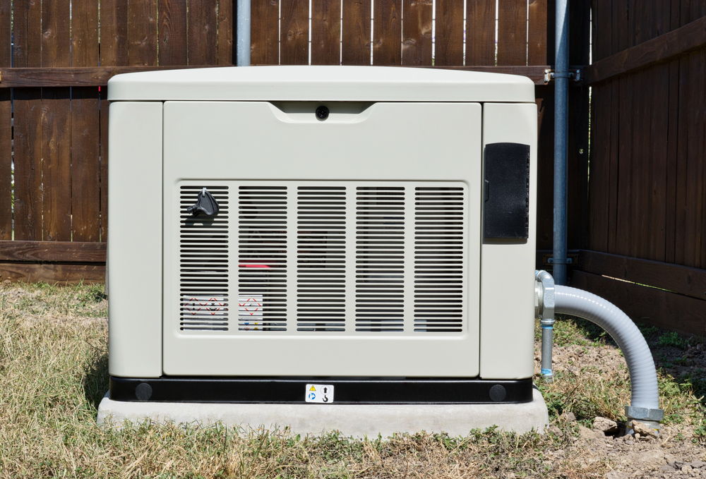 A standby home generator installation is set outdoors on a concrete pad, featuring a vented front panel and conduit attached on the right side, positioned beside a wooden privacy fence.