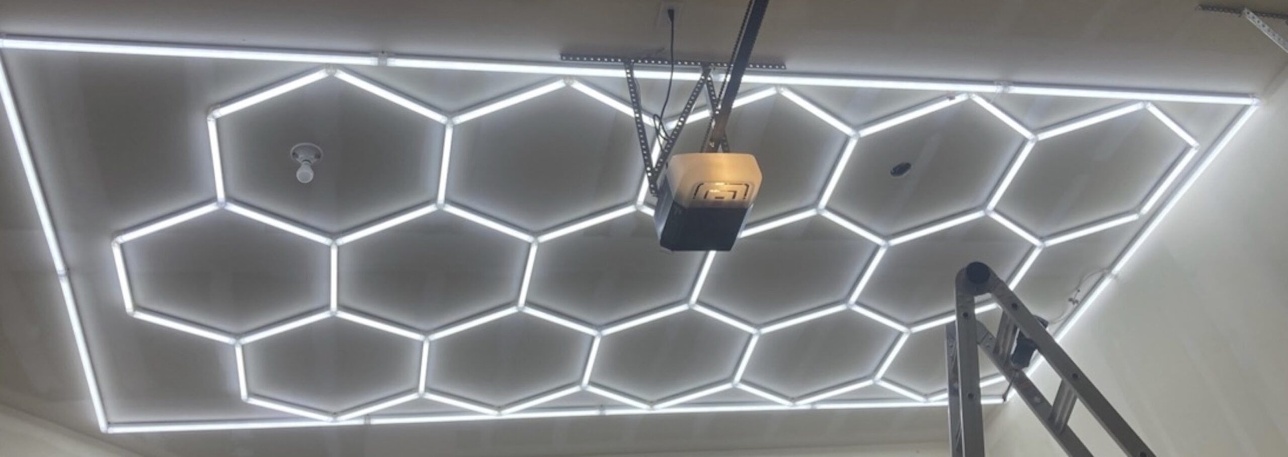 Hexagonal LED light panels are mounted on a ceiling near a garage door opener, with part of a ladder visible in the lower right corner.